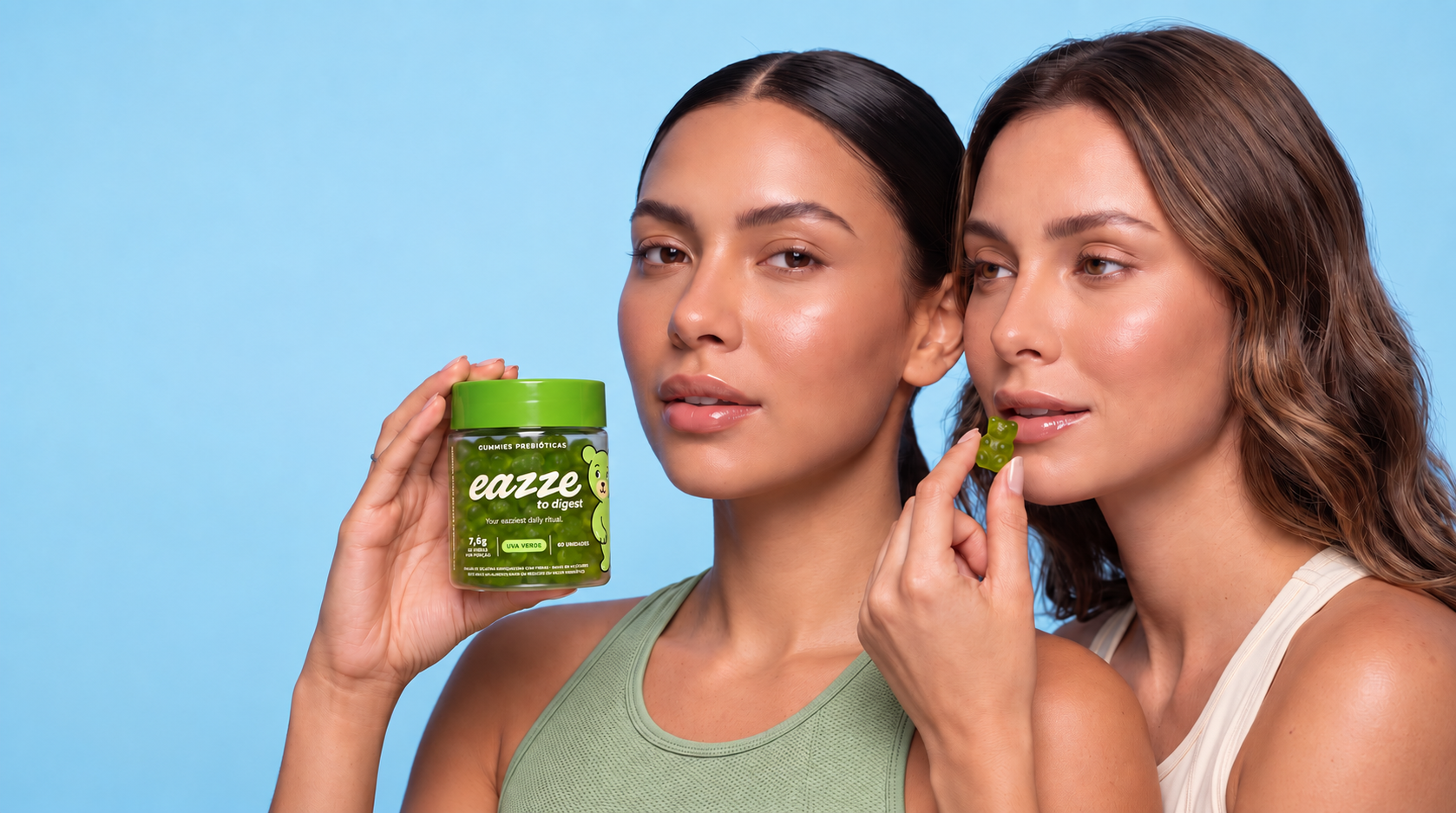 Duas mulheres juntas em fundo azul-claro: uma segura o pote completo EAZZE to Digest e a outra leva uma gummy verde aos lábios.